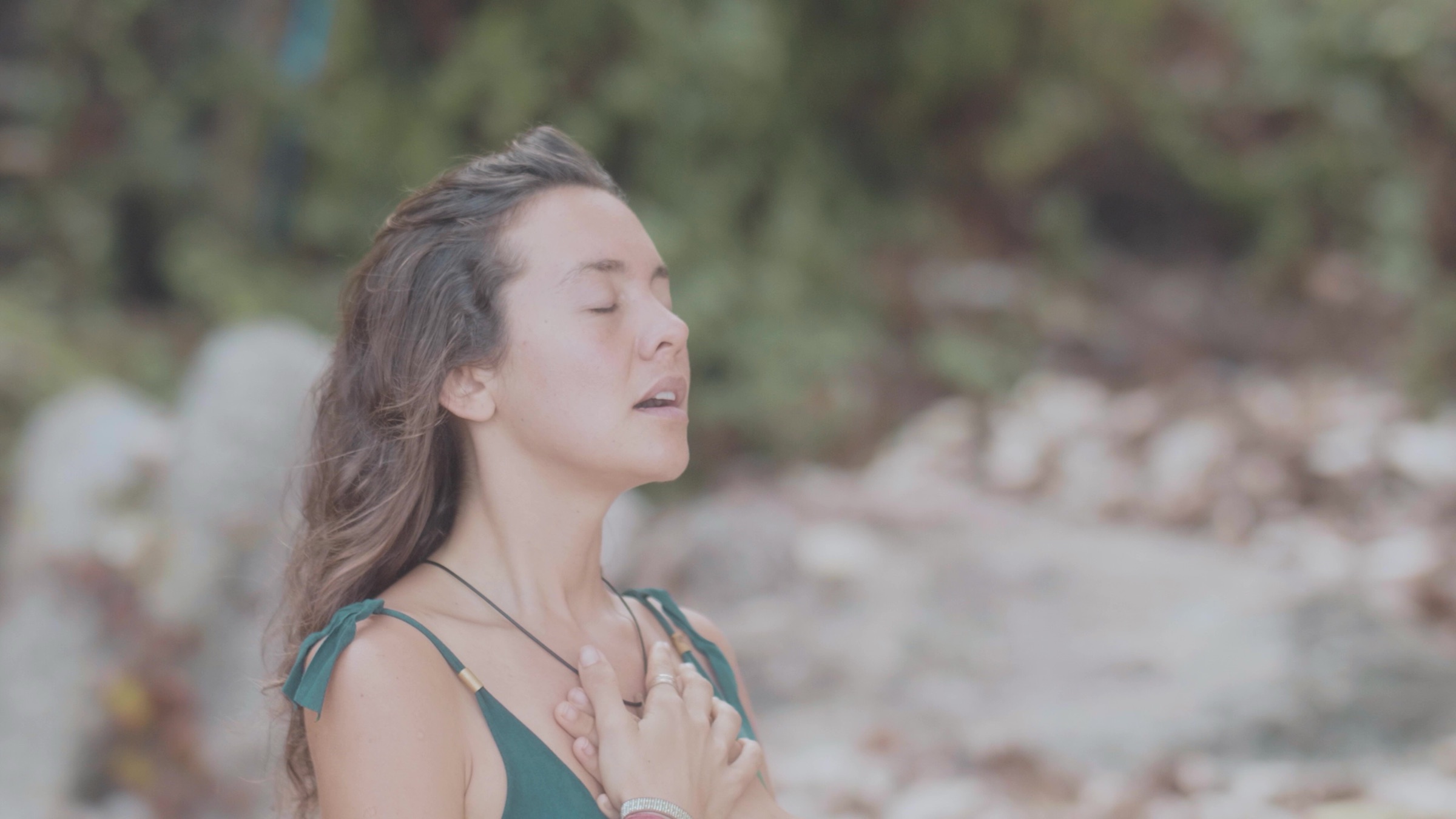 A woman with her hand on her heart, breathing, eyes closed, outdoors in soft natural light
