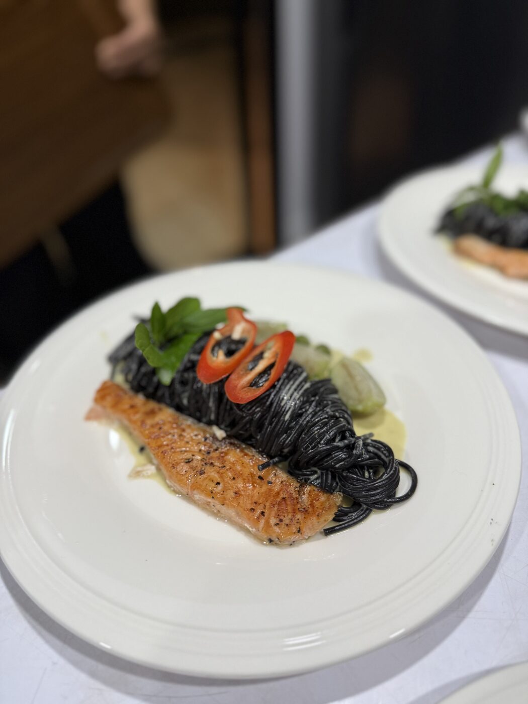 A plated dinner of seared salmon over black noodles with fresh herbs and chili