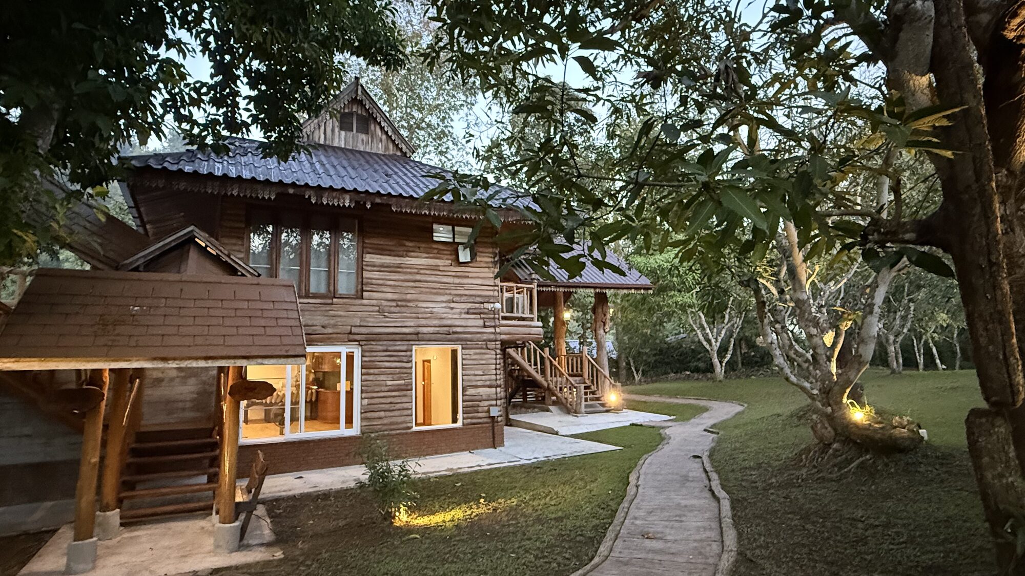 A two-storey wooden Lanna lodge lit warmly at dusk, a stone path leading through the garden